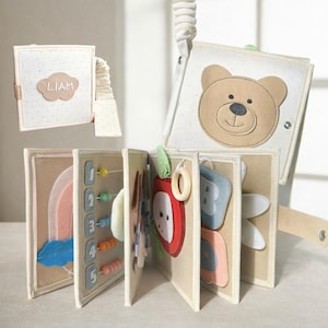 May include: A fabric quiet book featuring a beige cover with a brown bear face and a white cover with a cloud patch labeled LIAM. The interior pages include felt appliques of a rainbow, counting beads, an apple, and letters in soft colors.