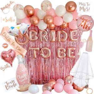 May include: A collection of party decorations for a bridal shower. Includes rose gold foil balloons spelling "Bride To Be", pink and white balloons, a sash, a heart-shaped balloon, and a veil. The backdrop is a pink metallic fringe.