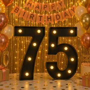 May include: Two large, black, three-dimensional marquee-style numbers forming 75, each illuminated by multiple warm white circular LED bulbs. The numbers are positioned on a surface with gold confetti, set against a backdrop of gold balloons.