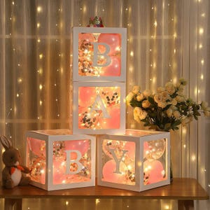 May include: White boxes spelling "BABY" with pink balloons, confetti, and string lights. Each box has a large letter cutout. A stuffed bunny and a crown are also visible.