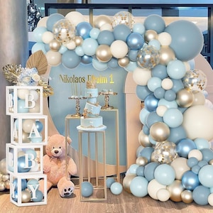 May include: A baby shower or birthday party decoration featuring a balloon arch in shades of blue, white, and gold. The scene includes a cake, a teddy bear, and boxes spelling "BABY". The backdrop has the text "Nikolaos Realm 1".