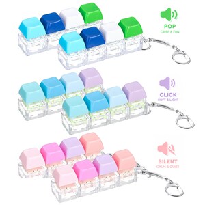 May include: Three sets of mechanical keyboard switch keychains with clear plastic bases and colorful keycaps. The top set features blue, white, and green caps with the text POP CRISP & FUN. The middle set has blue and purple caps with CLICK SOFT & LIGHT. The bottom set has pink and purple caps with SILENT CALM & QUIET.