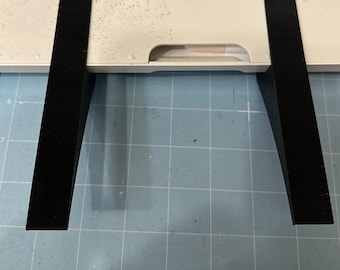May include: Two black, rectangular pieces of material are positioned on a white crafting machine. The pieces are angled, creating a V-shape. The machine sits on a blue grid surface.
