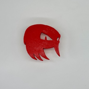 May include: A bright red, stylized wooden carving of a character's face, featuring a prominent eye and distinctive hair and spikes. The carving is set against a plain white background.