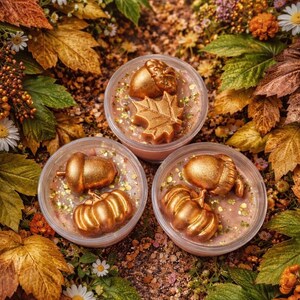 May include: Three clear containers filled with a light brown substance, each topped with gold-coloured autumn-themed decorations. The decorations include acorns, pumpkins, and a maple leaf, surrounded by glitter and set against a backdrop of fall foliage.