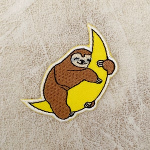 以下が含まれることがあります： 茶色のナマケモノが黄色の三日月をハグしている刺繍パッチ。