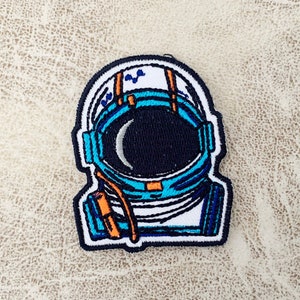 May include: An embroidered patch of an astronaut wearing a blue and white spacesuit with a black visor.