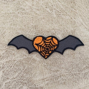 Puede incluir: Un murciélago negro con alas en forma de corazón naranja con un diseño de telaraña. El murciélago está bordado en una tela marrón claro.