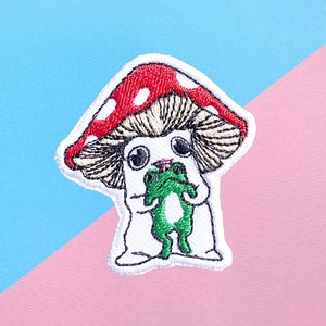 May include: Embroidered patch of a white mushroom character holding a green frog. The mushroom has a red cap with white dots and a white stem.