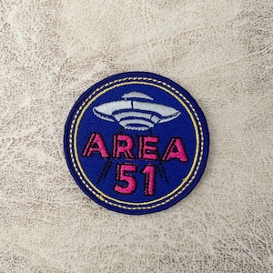 Puede incluir: Un parche bordado azul y amarillo con un OVNI blanco y el texto "AREA 51" en rosa.