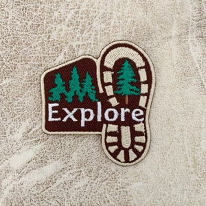 May include: Brown and white embroidered patch with a footprint design and the word "Explore" in white. The footprint has a green pine tree inside.