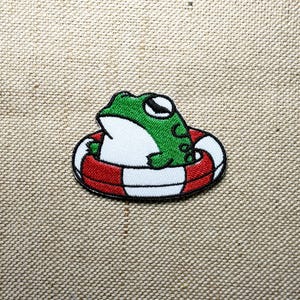 May include: Embroidered patch featuring a cartoon frog in a red and white striped life preserver. The frog is green and white with black eyes. The patch has a black border and is on a tan fabric background.