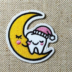 Könnte beinhalten: Gestickter Aufnäher mit einem lächelnden Zahn, der eine rosa-weiße Schlafhaube trägt, eingebettet in einen gelben Mond mit einem lächelnden Gesicht. Der Aufnäher hat einen weißen Rand und ist für Kleidung oder Accessoires bestimmt.