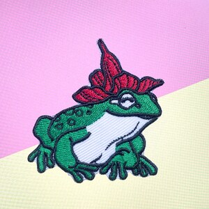 May include: Green embroidered frog with a red flower on its head. The frog is sitting on a light yellow surface.