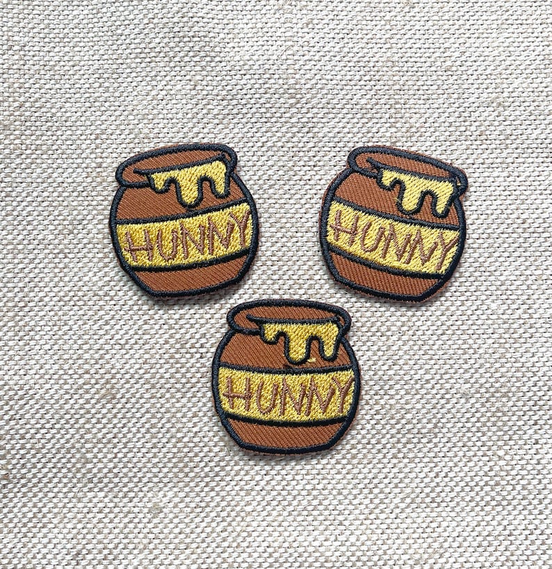 May include: Three embroidered iron-on patches in the shape of honey pots with the word "Hunny" written on them. The patches are brown with yellow honey dripping from the top.