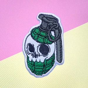 May include: Embroidered patch depicting a skull-shaped grenade. The patch features a white skull with black eye sockets and a green plaid pattern. The grenade's top is grey with a pull ring, all outlined in white.