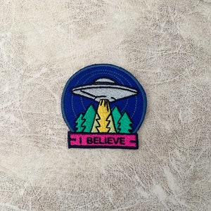 May include: A blue and white embroidered patch with a UFO flying over a forest and the text "I BELIEVE".
