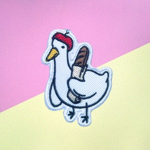 May include: Embroidered patch of a white goose wearing a red beret and holding a baguette. The goose is outlined in blue thread.