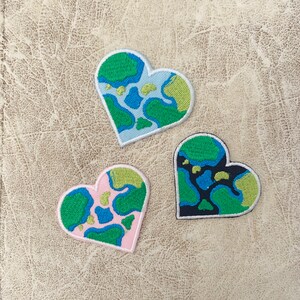 May include: Three heart-shaped patches with a world map design. The patches are embroidered with green, blue, and black thread on a white background. The hearts are different sizes and are arranged in a triangular shape.