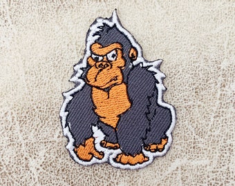 Può includere: Una toppa ricamata con un gorilla dei cartoni animati con un'espressione seria, in tonalità di grigio e arancione.