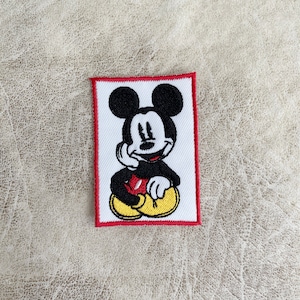 Könnte beinhalten: Ein gestickter Aufnäher mit rotem Rahmen, der Mickey Mouse in sitzender Pose mit einem rot-gelben Farbschema zeigt.