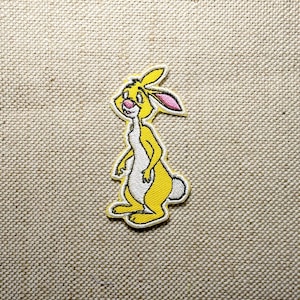 May include: A yellow and white embroidered patch featuring a cartoon rabbit with pink inner ears. The rabbit is standing upright with a white belly and a small white tail. The patch has a yellow border and is set against a textured beige background.