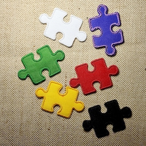 Könnte beinhalten: Sechs farbige Puzzleteile in Weiß, Grün, Lila, Rot, Gelb und Schwarz. Jedes Teil hat einen gestickten Rand und ist auf einer strukturierten, beigefarbenen Stoffoberfläche angeordnet.