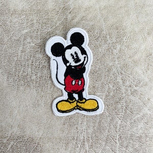May include: A black and white embroidered patch of Mickey Mouse with red shorts and yellow shoes. The patch is on a light brown fabric background.