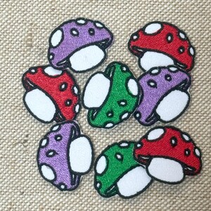 May include: Embroidered mushroom patches in red, purple, and green with white spots and white undersides. The patches have a black outline and are arranged on a tan fabric background. These patches are suitable for clothing and accessories.