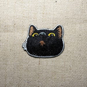 Op de afbeelding: Geborduurde patch met de kop van een zwarte kat met gele ogen en bruine oren en neus. De patch heeft een witte rand en is ontworpen om op stof te strijken of te naaien. Het gezicht van de kat is gecentreerd.