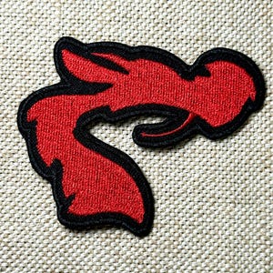 May include: Embroidered patch featuring a stylized red flame design with a black outline. The patch has a textured appearance, likely made from fabric or thread. The design is set against a neutral, woven background.