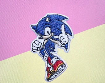 May include: Embroidered patch featuring Sonic the Hedgehog in a dynamic pose. The patch showcases Sonic's blue fur, white gloves, and red shoes with yellow accents. The patch is outlined in white and set against a pink and yellow background.