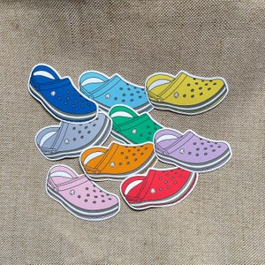 Puede incluir: Nueve pares de zuecos coloridos en varios tonos de azul, verde, amarillo, naranja, rojo y morado. Los zuecos están hechos de un material similar al caucho y tienen un diseño clásico de zueco.