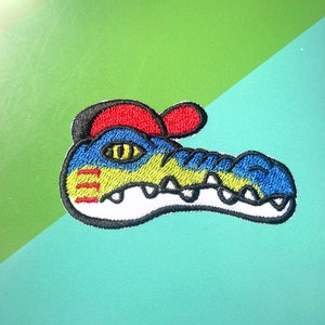 May include: Embroidered patch of a blue, yellow and white alligator wearing a red baseball cap. The alligator has a toothy grin and a yellow eye.