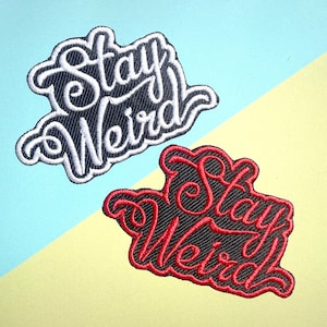 Pode incluir: Dois patches bordados com o texto "Stay Weird" em contorno branco sobre fundo preto. O patch superior tem texto branco e o patch inferior tem texto vermelho.