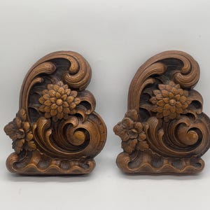 May include: Two ornate, wooden decorative wall appliques. Each features a central flower, smaller flowers, and swirling, carved details. The appliques are a warm brown color and have a vintage aesthetic.