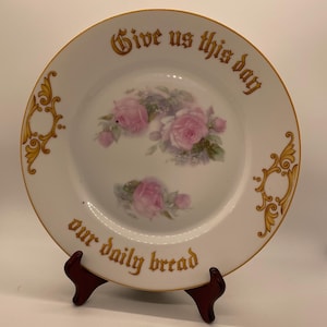 May include: White porcelain plate with a gold rim and floral rose design. The plate features the text "Give us this day our daily bread" in gold lettering. Decorative gold flourishes are on the plate's edge.