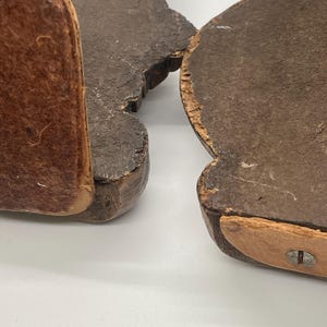May include: Two dark brown wooden bookends with a distressed finish. Each bookend has a curved shape with a leather-like material attached to the bottom and side for support. The bookends are set against a white background.