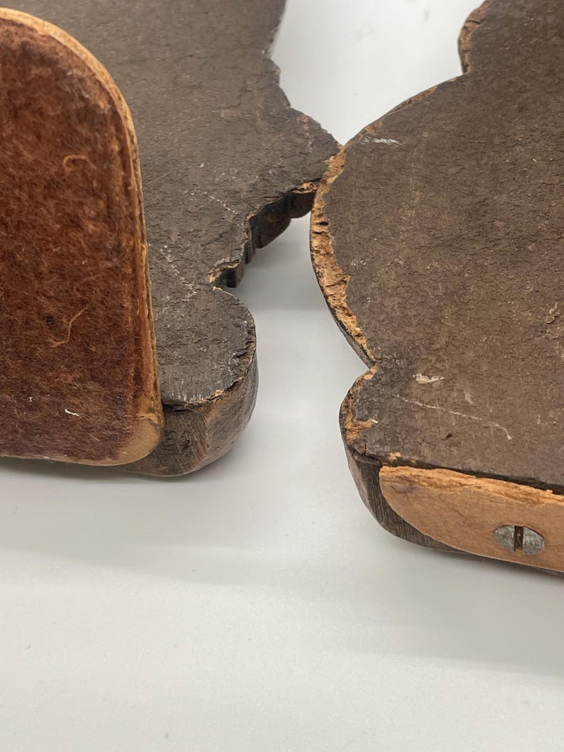 May include: Two dark brown wooden bookends with a distressed finish. Each bookend has a curved shape with a leather-like material attached to the bottom and side for support. The bookends are set against a white background.