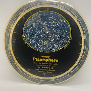 May include: A vintage Philips Planisphere, a circular astronomical instrument. The black face displays star charts and constellations, with text indicating visibility for Latitude 42°N. The outer ring features months and dates. The text reads "Philips' Planisphere".