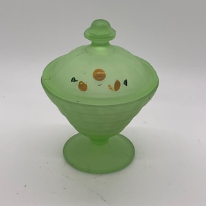 May include: A green glass candy dish with a lid. The dish has a pedestal base and a ribbed bowl. The lid is decorated with small, painted gold and black accents. The candy dish is a vintage piece.
