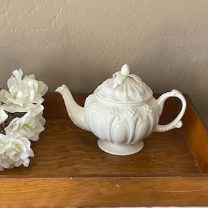 May include: A white ceramic teapot with a decorative lid and handle sits on a wooden tray. The teapot has a detailed design and is next to a basket of white flowers. The tray is brown.