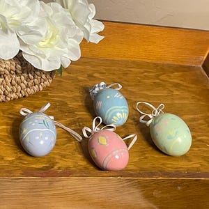 May include: Four decorative Easter eggs in pastel colors with floral designs and ribbon hangers. The eggs are light blue, pink, and green, arranged on a wooden surface. A basket of white flowers is in the background.
