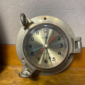May include: A round, brass-colored quartz clock with a nautical design. The clock face is gold with black numerals and red second hand. The words "ROYAL MARINER" and "QUARTZ" are printed on the face. The clock has two ring handles.