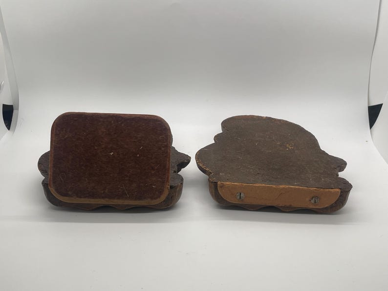 May include: Two brown wooden bookends with a decorative design. One bookend has a brown velvet backing, while the other shows the underside with a wooden base and screws. The bookends are on a white surface.