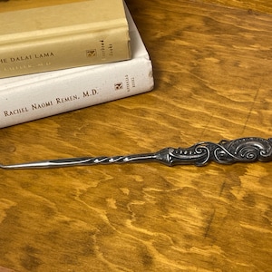 May include: A silver-toned metal button hook featuring a twisted shaft and an ornate, embossed handle with intricate scrollwork and decorative detailing. The tool has a curved hook at one end and a solid, sculpted grip at the opposite end.