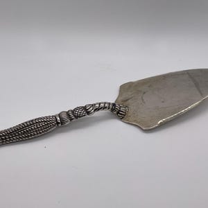 May include: Silver-toned cake server with a decorative handle. The handle features a woven rope design, transitioning to a twisted rope detail near the blade. The blade has a rounded, slightly curved shape.