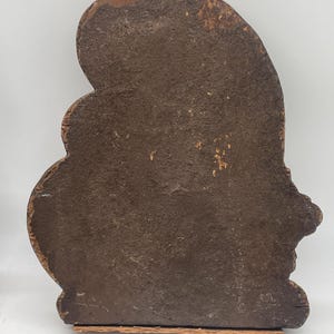 May include: A weathered, wooden decorative piece with a dark brown, textured surface. The object has an irregular, organic shape with a rounded top and a flat base. The wood shows signs of age and wear.