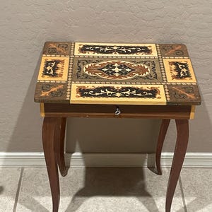 May include: A rectangular wooden side table with four curved legs and a single drawer featuring a small silver knob. The tabletop displays an intricate inlaid marquetry design in shades of cream, dark brown, and tan with scrolling floral motifs.
