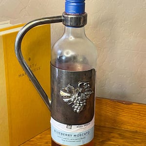 May include: A clear glass bottle of Blueberry Moscato with a blue screw cap and a silver-toned metal holder featuring a grape cluster emblem. The label reads Blueberry Moscato, light and spritzy, with refreshing real blueberry juice and real fruit.